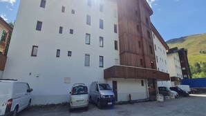 Un edificio de varios pisos con fachada blanca y una sección de madera a la derecha. Hay varios coches aparcados en la calle.