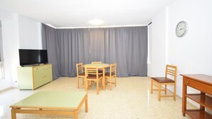 Une pièce avec un téléviseur posé sur un meuble, une table à manger avec des chaises, une chaise en bois et une horloge au mur.