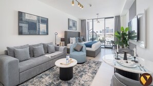 A modern hotel room with a grey sofa, a round coffee table, a dining area with a table set for two, and a large window offering a city view. - Vogue | Binghatti Galaxy JVC near Circle Mall (Dubai)