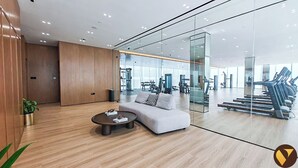 A modern hotel lobby with a wooden reception desk, a sofa, a coffee table, and a glass wall leading to a gym area with exercise equipment. - Vogue | Binghatti Galaxy JVC near Circle Mall (Dubai)