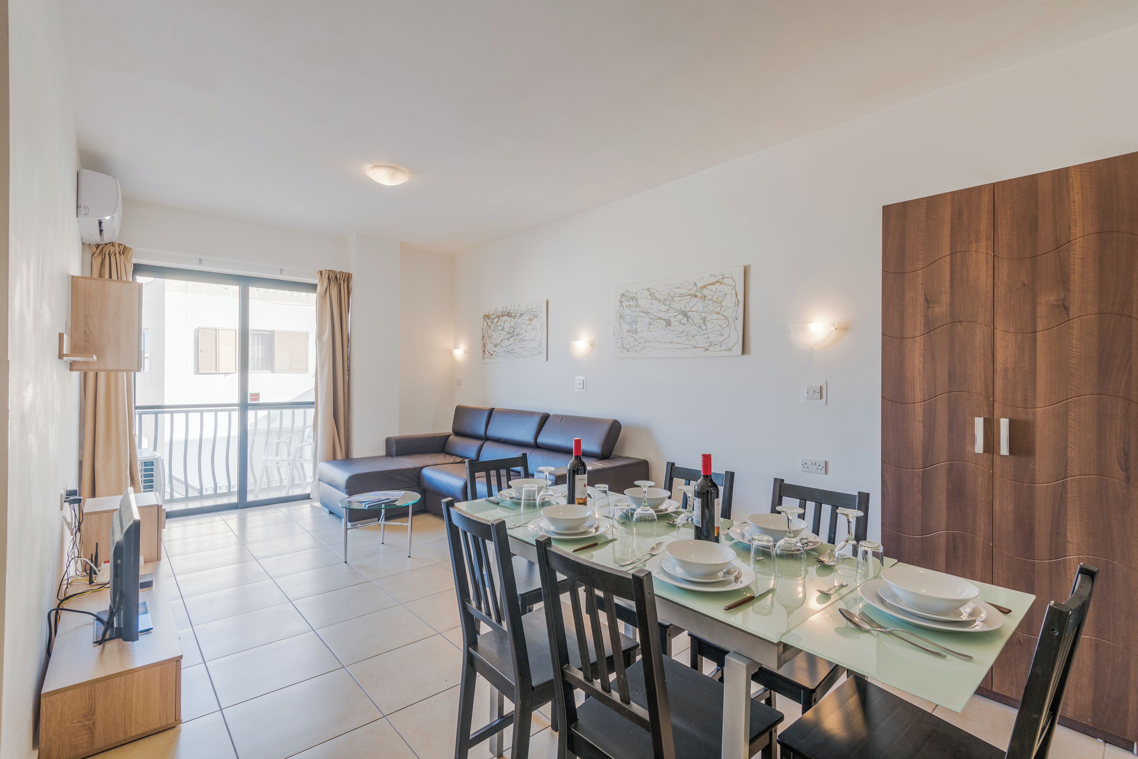 A dining area with a table set for four, a sofa, and a balcony with curtains.