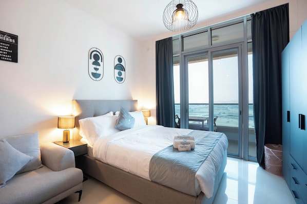 A hotel room with a bed, a sofa, a nightstand, a lamp, and a view of the sea through large windows. - Bloomfields Studio in Julphar Residence-Reem (Abu Dhabi)