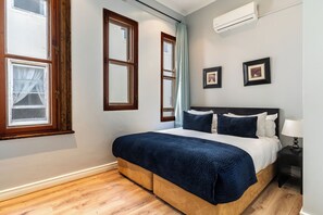 A bedroom with a bed, wooden flooring, a window with curtains, and a wall-mounted air conditioner.