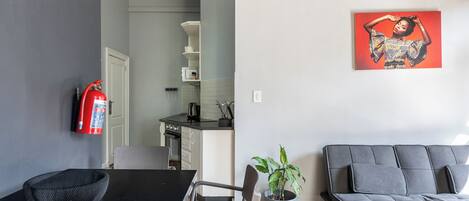 A modern living room with a grey sofa, a dining table with chairs, a potted plant, and a fire extinguisher on the wall.