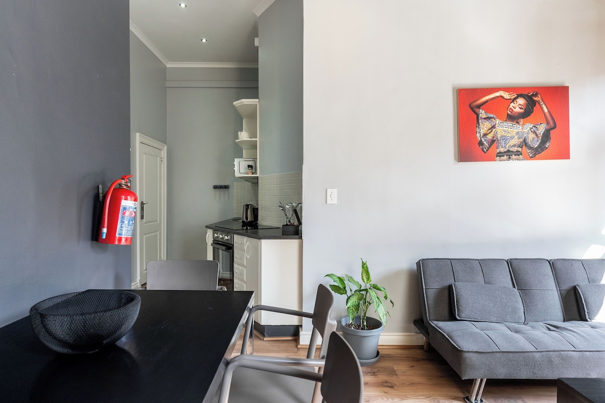 A modern living room with a grey sofa, a dining table with chairs, a potted plant, and a fire extinguisher on the wall.
