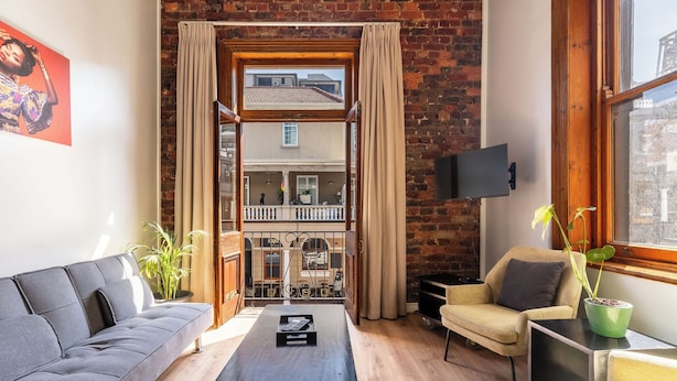 A modern living room with a sofa, a coffee table, a TV, and a view of a balcony and building through large windows. - Long Street Self Catering Apartments (Cape Town)