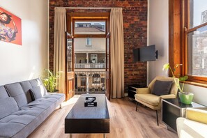 A modern living room with a sofa, a coffee table, a TV, and a view of a balcony and building through large windows.