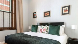 A neatly made bed with a dark green blanket, a wooden headboard, and green and floral pillows. There are framed pictures on the wall and a window with bars. - Long Street Self Catering Apartments (Cape Town)