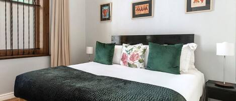 A neatly made bed with a dark green blanket, a wooden headboard, and green and floral pillows. There are framed pictures on the wall and a window with bars.