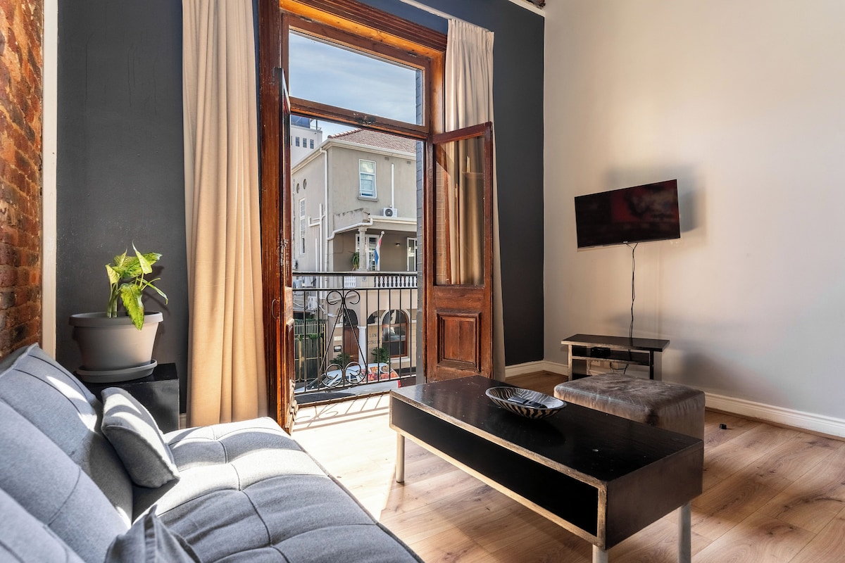 A modern living room with a sofa, a coffee table, a TV, and a view of a balcony with a building in the background.