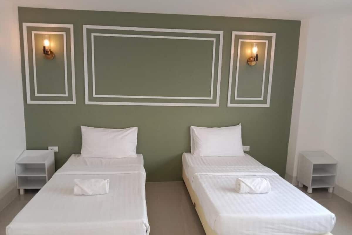 A room with two beds, each with white linens and pillows, against a green wall with decorative lighting fixtures.