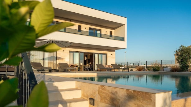A modern two-story building with a swimming pool, outdoor seating, and a view of the sea.