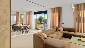 A modern living room with a sectional sofa, a dining area with a round table and chairs, and a large sliding glass door leading to a balcony with a view of the sea.