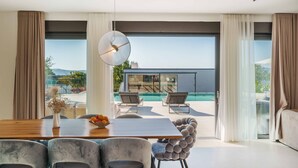 A modern living room with a large dining table, two lounge chairs, and a view of a swimming pool and outdoor area.