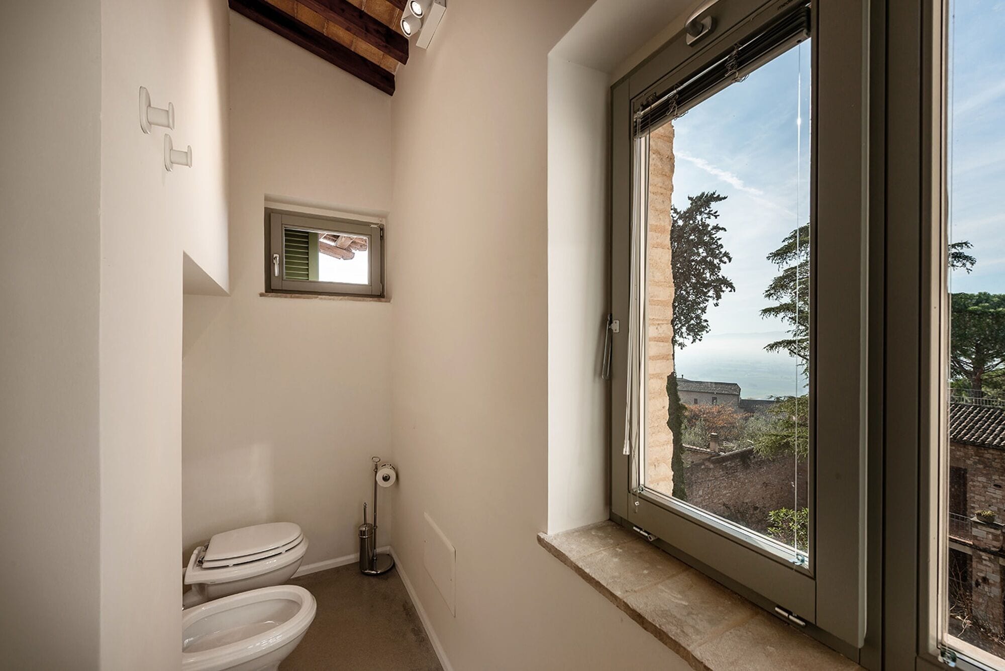 Une petite salle de bain avec des toilettes, une fenêtre et une vue sur les arbres et les bâtiments à l’extérieur.