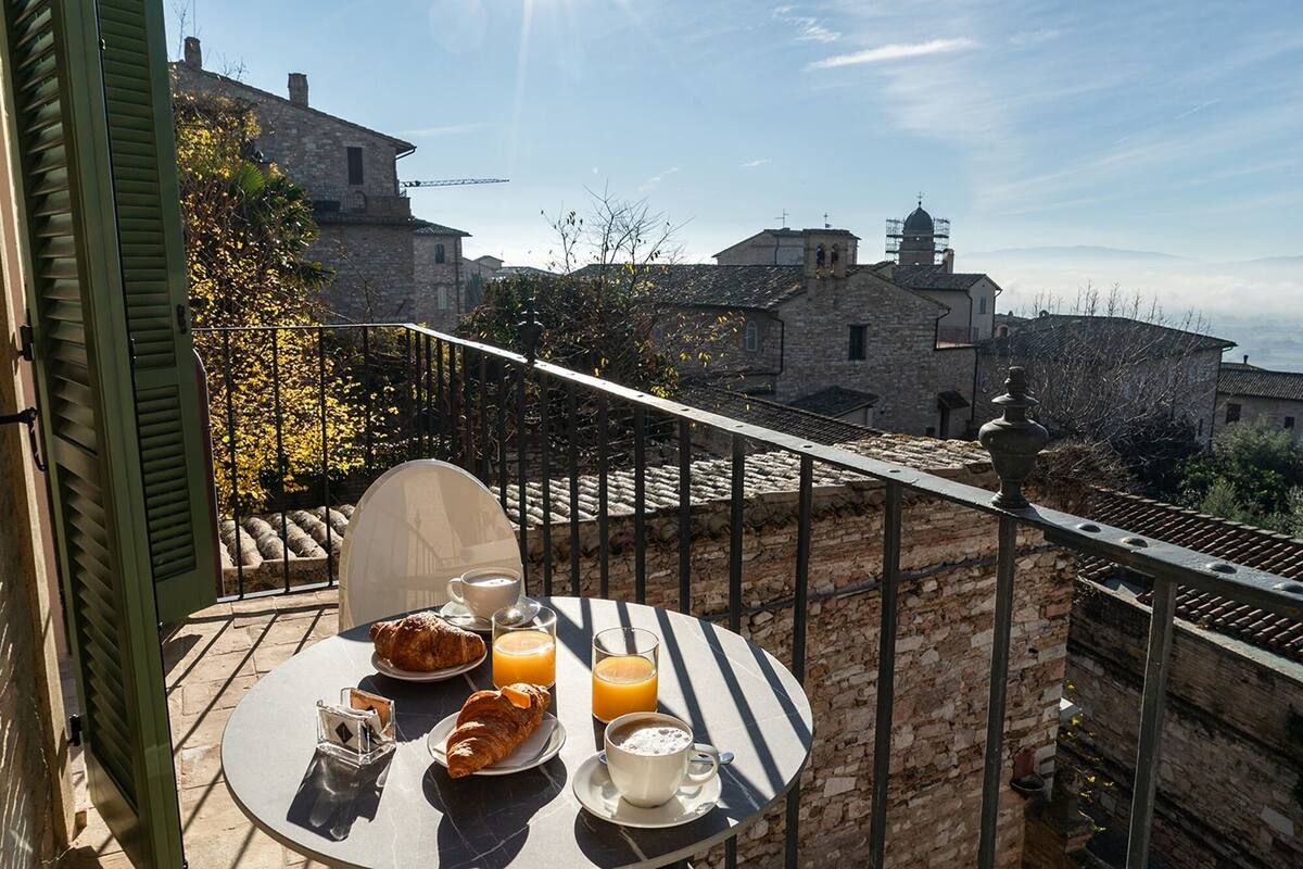 A table with croissants, orange juice, and coffee on a balcony overlooking historic stone buildings.