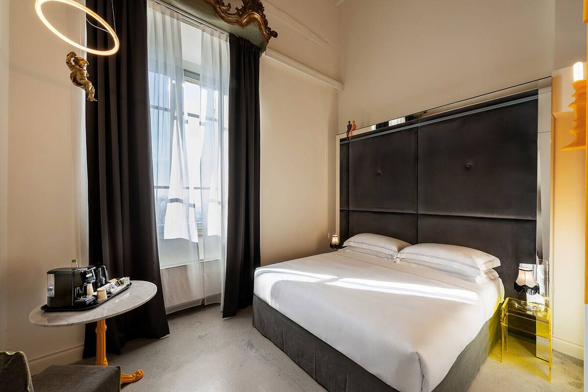 A modern hotel room with a large bed, a small round table with a coffee maker, a bedside lamp, and a view of the city through large windows.