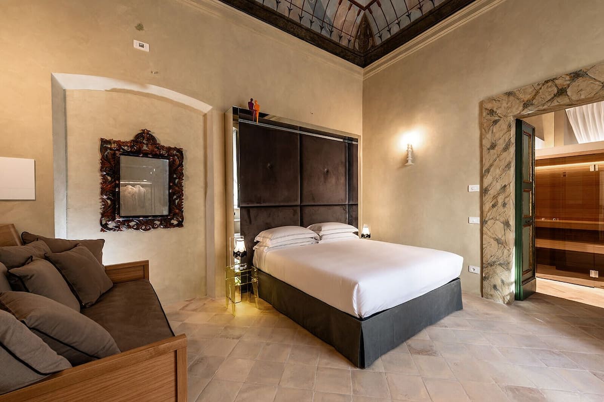 A bedroom with a large bed, a wooden bench, a mirror, and a nightstand with a lamp.