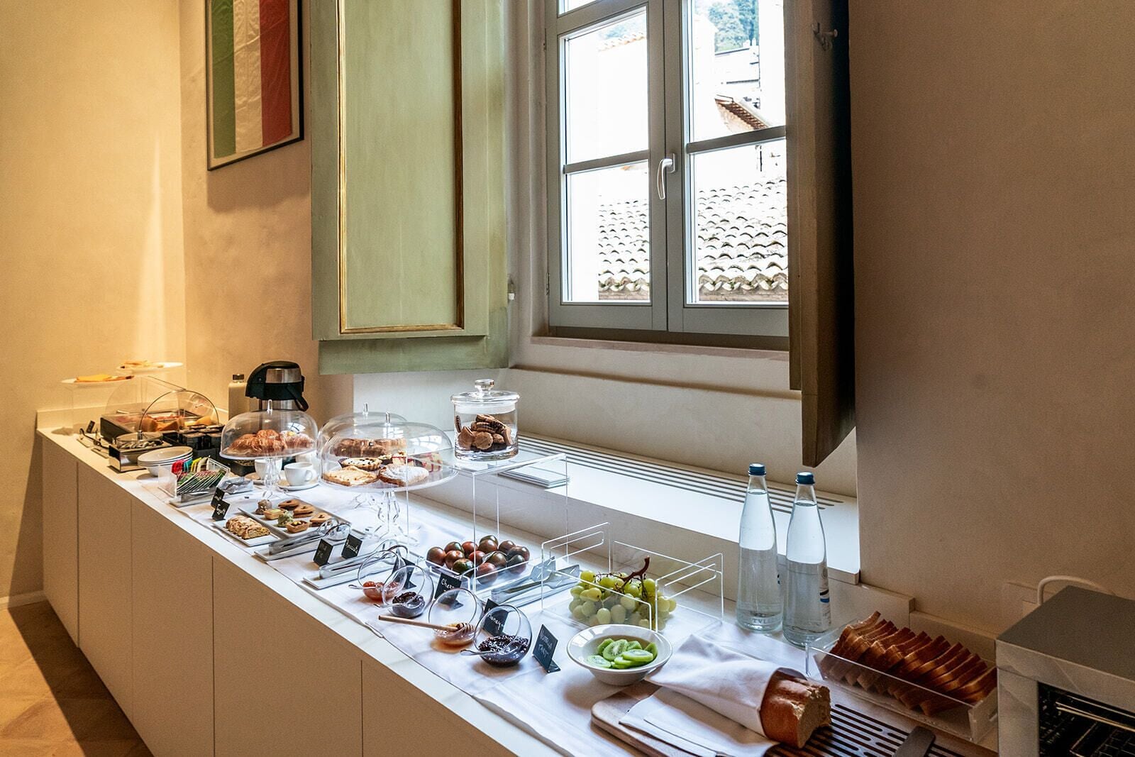 Een ontbijtbuffet met diverse gerechten en drankjes, een raam met uitzicht op een pannendak en een keuken met een broodpresentatie.