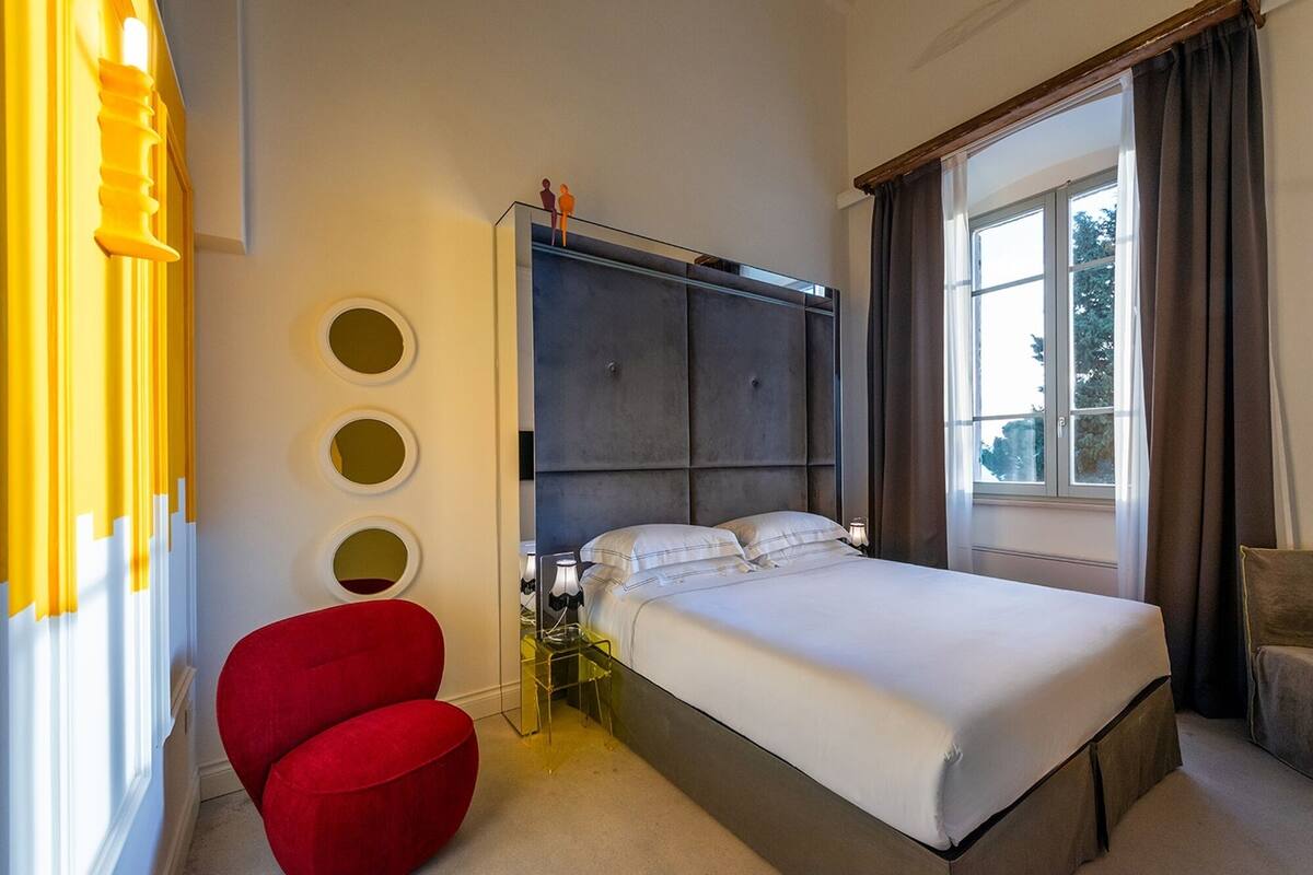 A bedroom with a large bed, a red chair, a glass table, and a window with curtains.