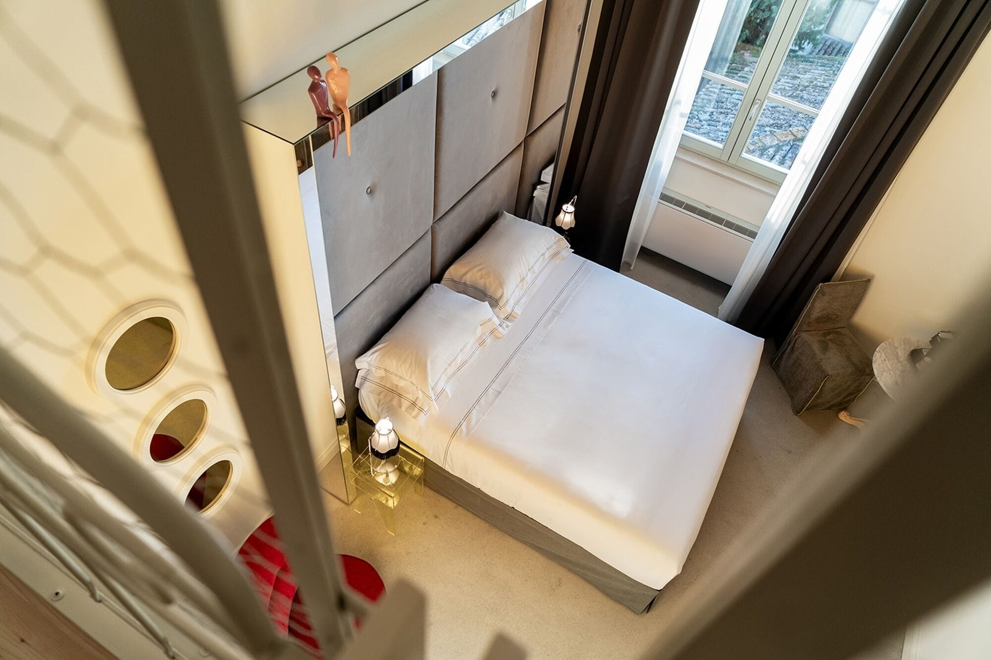 A modern hotel room with a large bed, a bedside table, a mirror, and a window with a view of the outside.