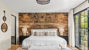 A bedroom with a stone wall, a large bed, two bedside tables, and a view through a glass door. - Palo Verde Experience|Zen-Inspired Resort Oasis (Phoenix)