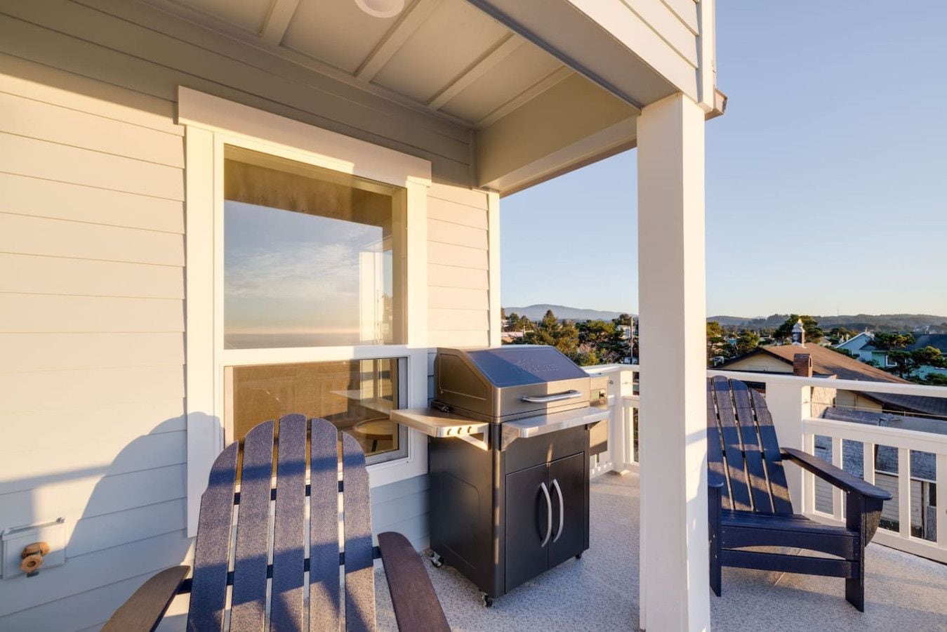 A covered outdoor area with a grill, Adirondack chairs, and a view of a residential neighborhood.