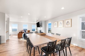 Un vaste espace dédié à la salle à manger, avec une longue table et des chaises, un coin salon confortable avec un canapé et des coussins, et une grande fenêtre offrant une vue sur l  ’océan.