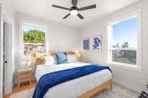 Une chambre à coucher comprenant un lit, une table de chevet, un ventilateur de plafond, deux fenêtres et des œuvres d’art accrochées au mur.