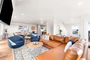 Una sala de estar moderna con un sofá de cuero marrón, dos sillones azules, una mesa de centro redonda y una alfombra estampada. Hay una zona de comedor con una mesa y sillas, y una cocina al fondo.
