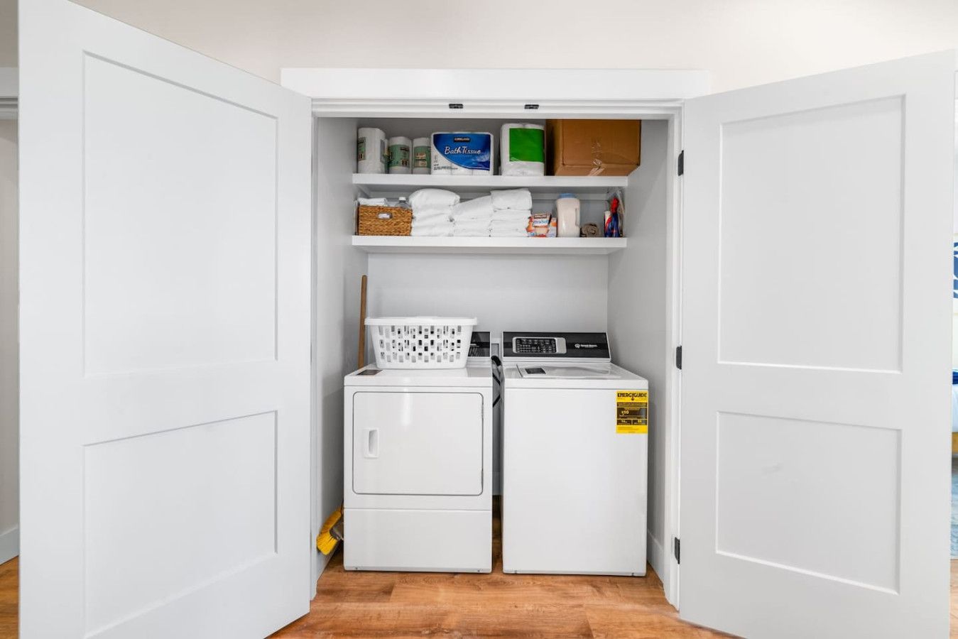 A compact laundry room with two stacked washing machines, a dryer, and shelves above containing various laundry supplies.