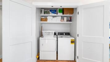 A compact laundry room with two stacked washing machines, a dryer, and shelves above containing various laundry supplies.