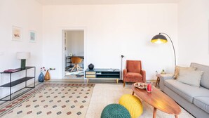 Una sala moderna con sofá, mesa de centro y lámpara. Hay una silla y una mesita con un jarrón y una lámpara. La habitación tiene una alfombra estampada y una puerta que da a otra estancia.