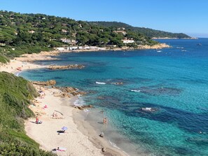 A coastal beach with clear turquoise waters, sandy shore, and a hillside with buildings.