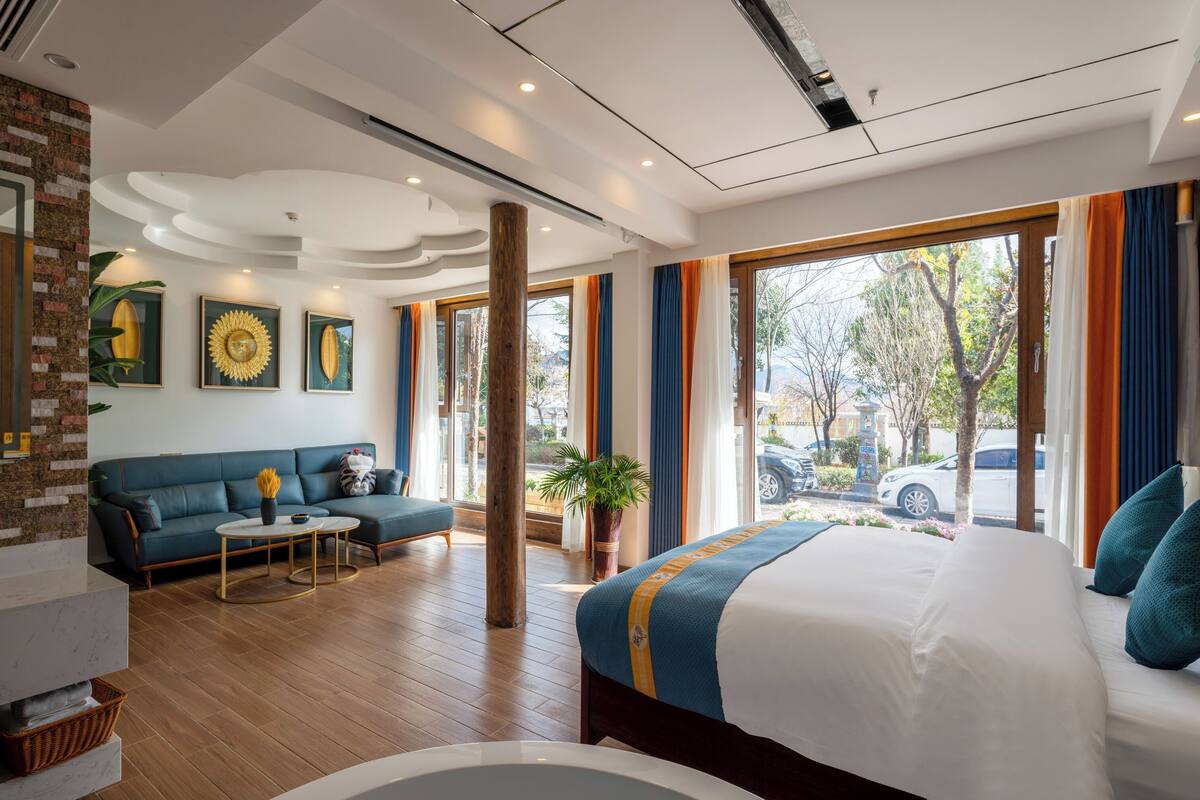 A modern hotel room with a large bed, a sofa, a small table, and a view of the street.