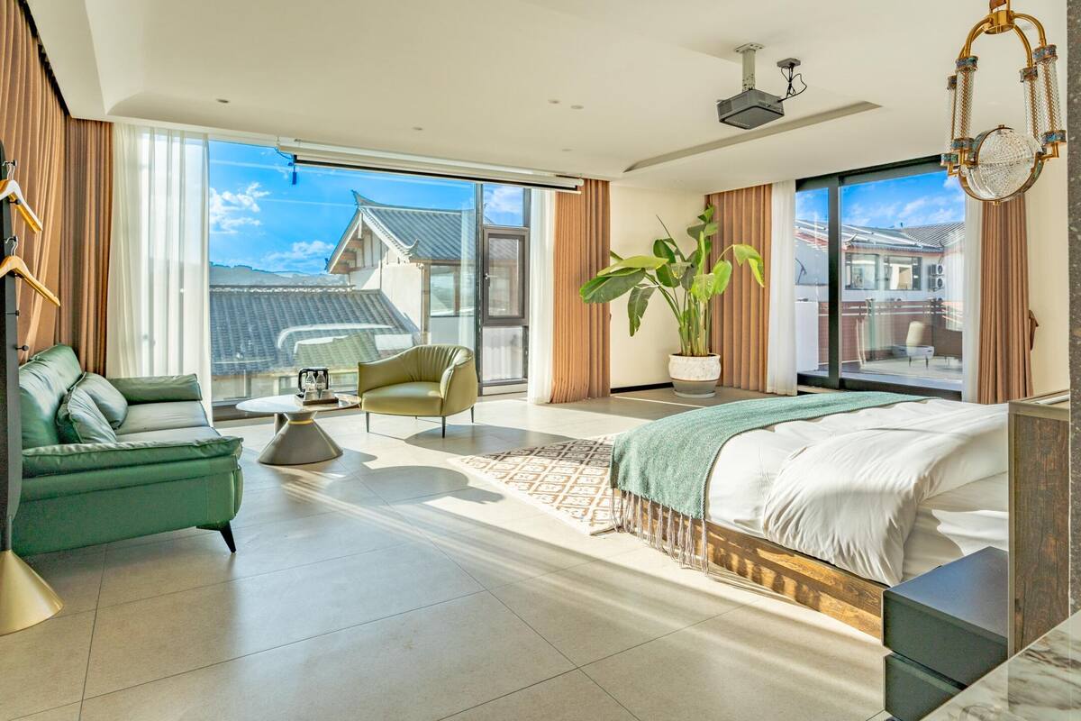 A modern hotel room with a large bed, a green armchair, a sofa, a potted plant, and a view of the city.
