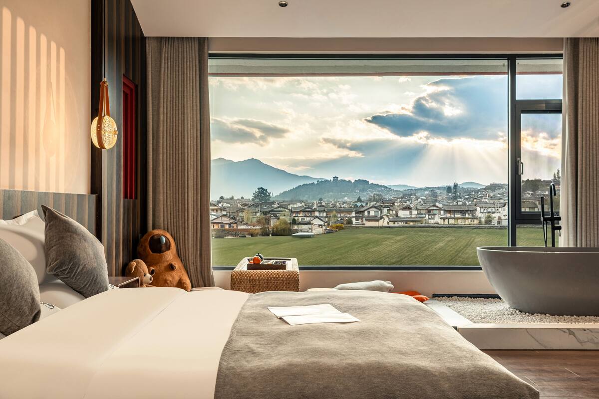 A modern hotel room with a large window offering a view of a cityscape and mountains.