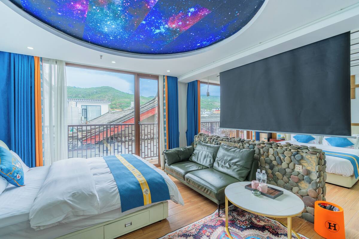 A modern hotel room with a large bed, a sofa, a small table, and a ceiling with a starry sky projection.