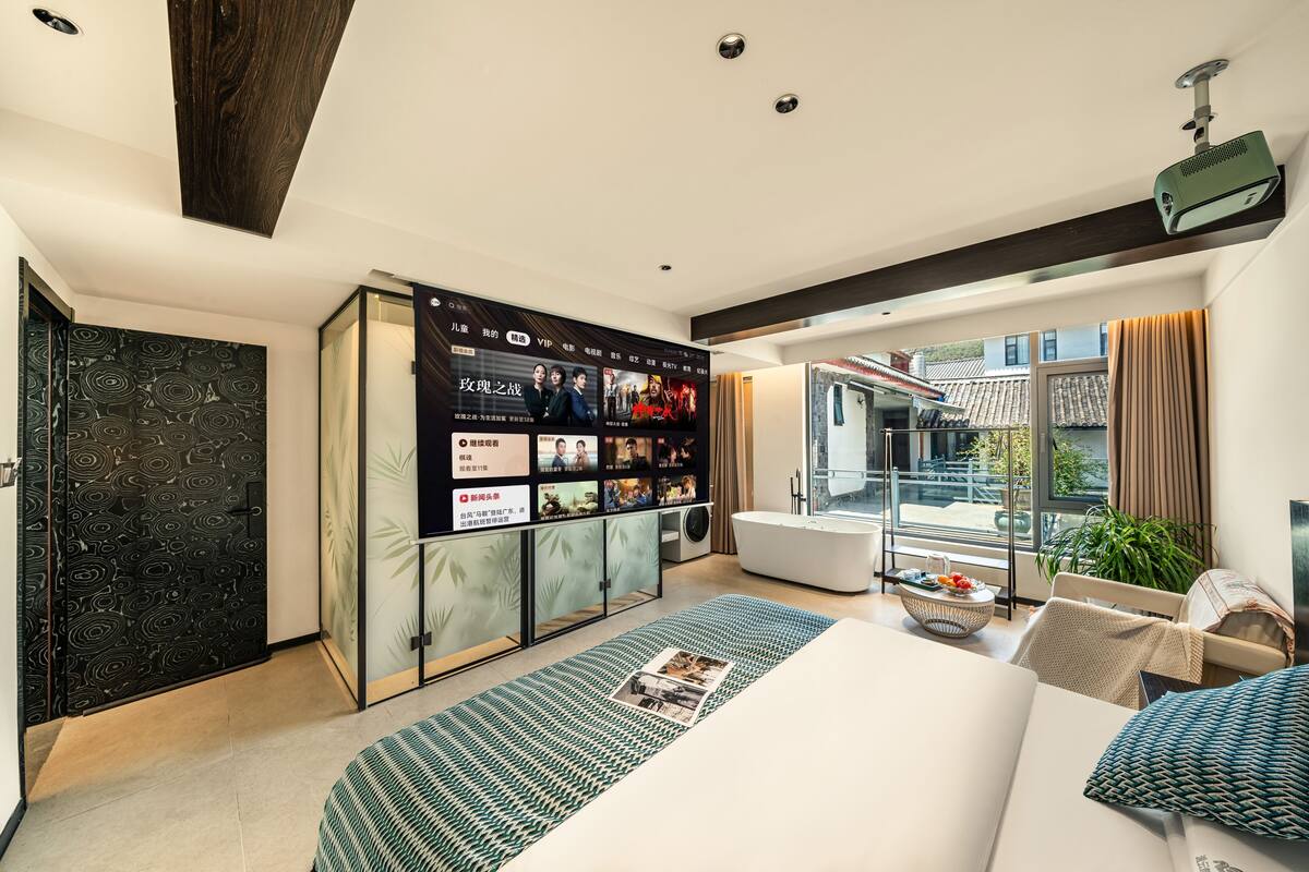A modern hotel room with a large bed, a seating area with a small table, a built-in entertainment system, and a balcony with a view of the city.