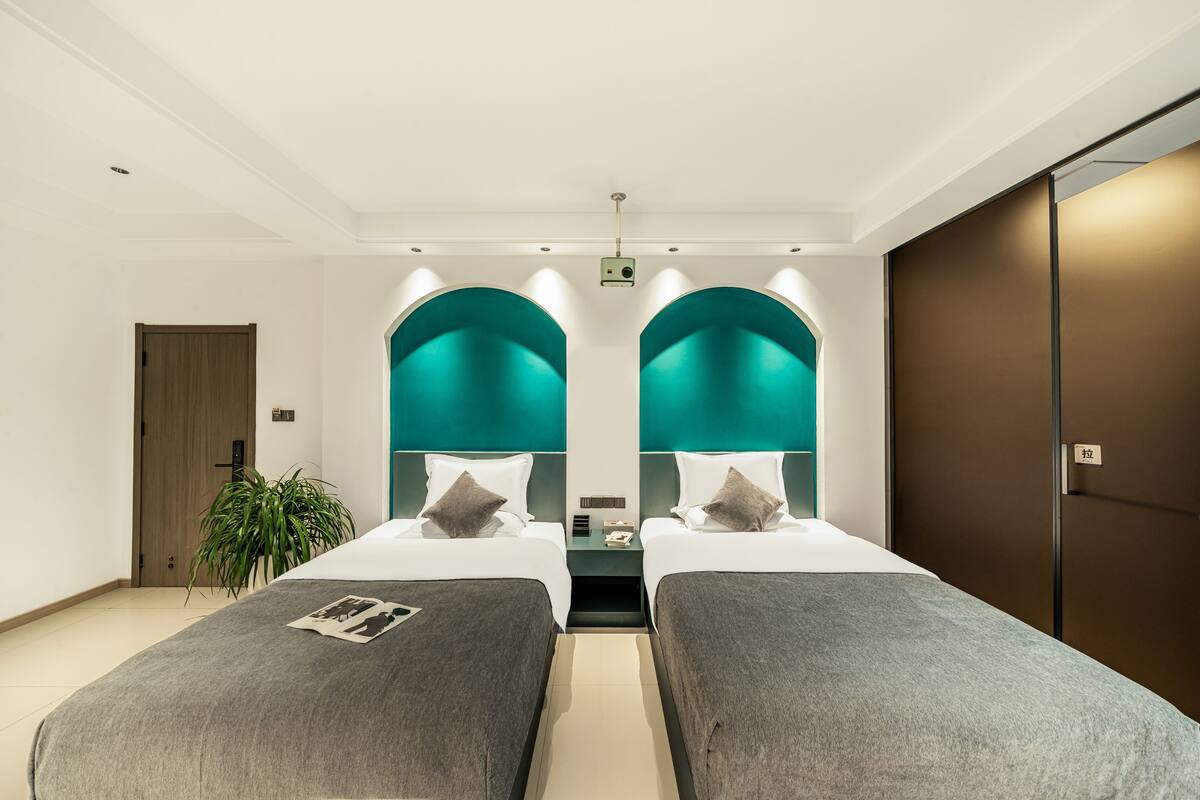 A modern hotel room with two single beds, a large mirror, a potted plant, and a wooden door.