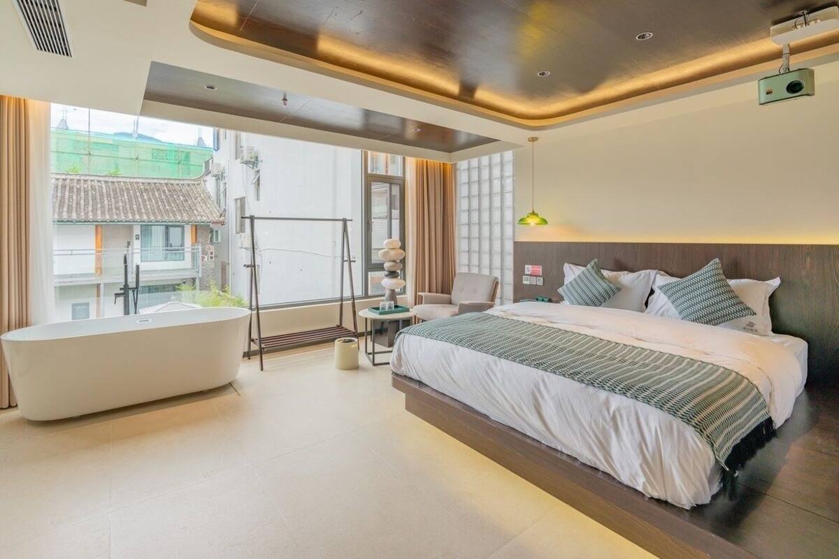 A modern hotel room with a large bed, a bathtub, and a balcony with a view of buildings.