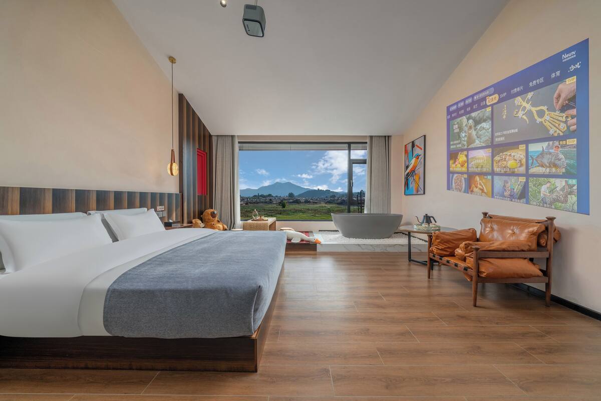 A modern hotel room with a large bed, a seating area with a sofa and armchair, a large window with a view of a mountain, and a wall with a menu board.