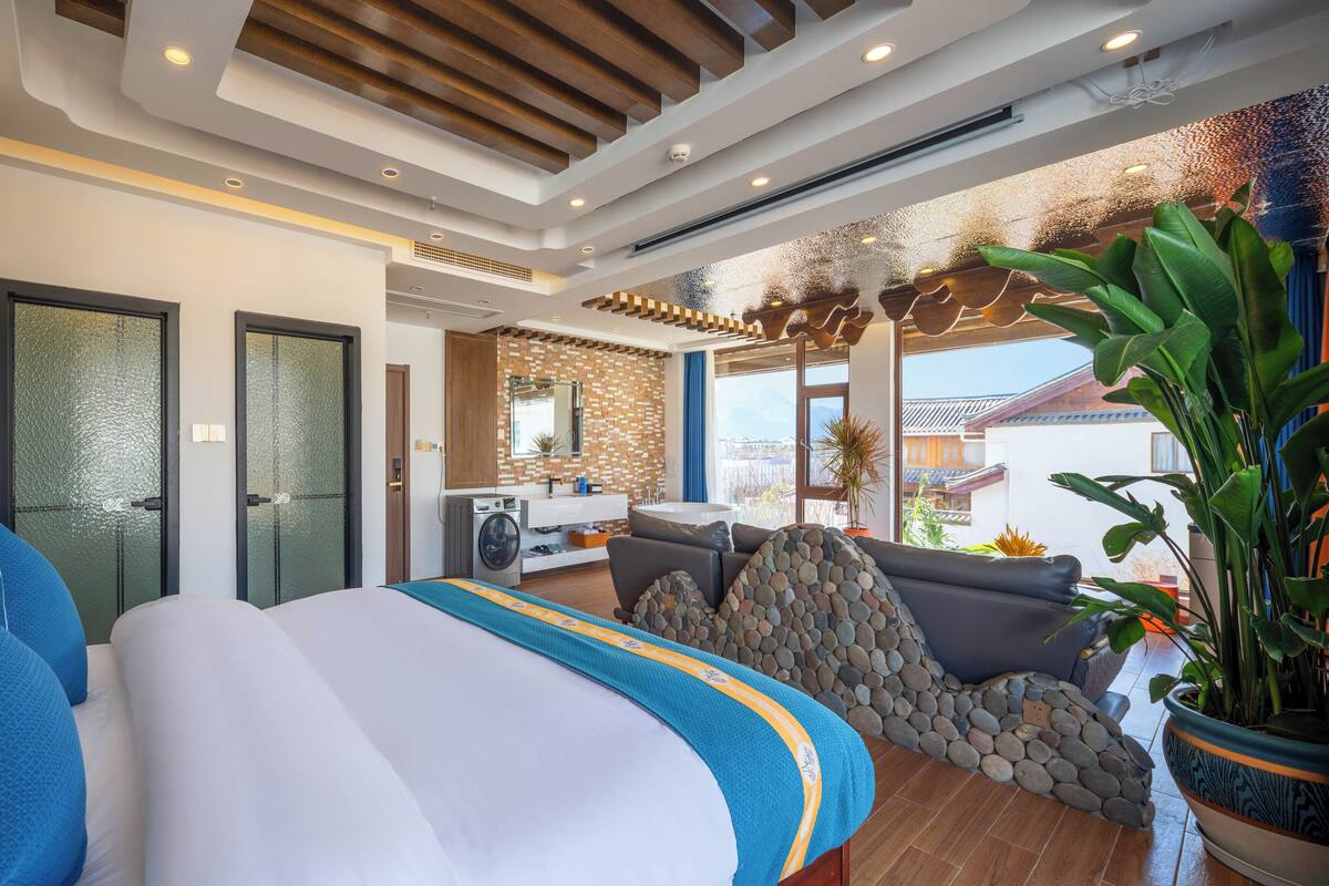 A modern hotel room with a large bed, a bathroom with a shower, a kitchen area, and a view of the outside.