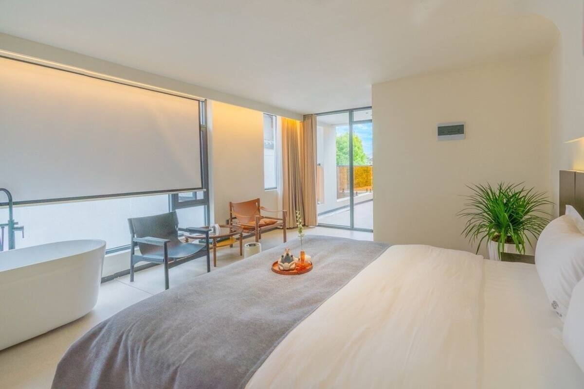 A modern hotel room with a large bed, a bathtub, a seating area with chairs, and a view of the outdoors.