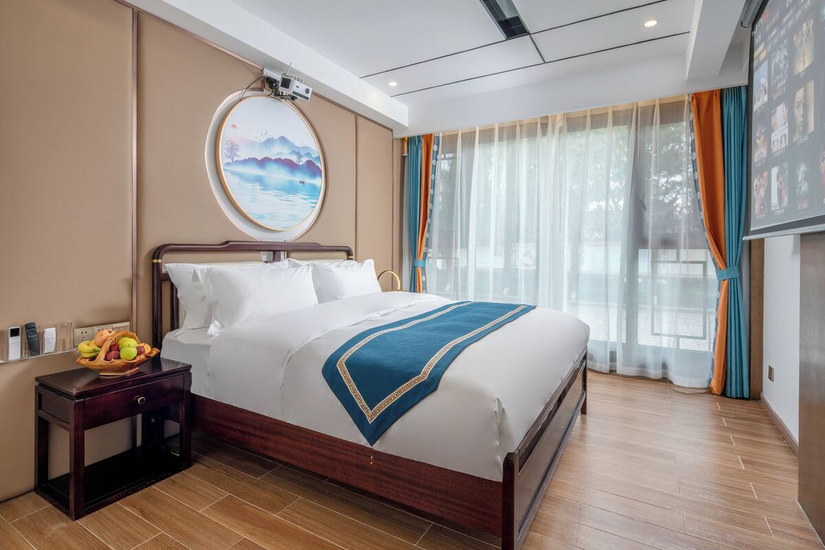 A hotel room with a bed, a bedside table with a fruit bowl, a wall-mounted TV, and a large window with sheer curtains.