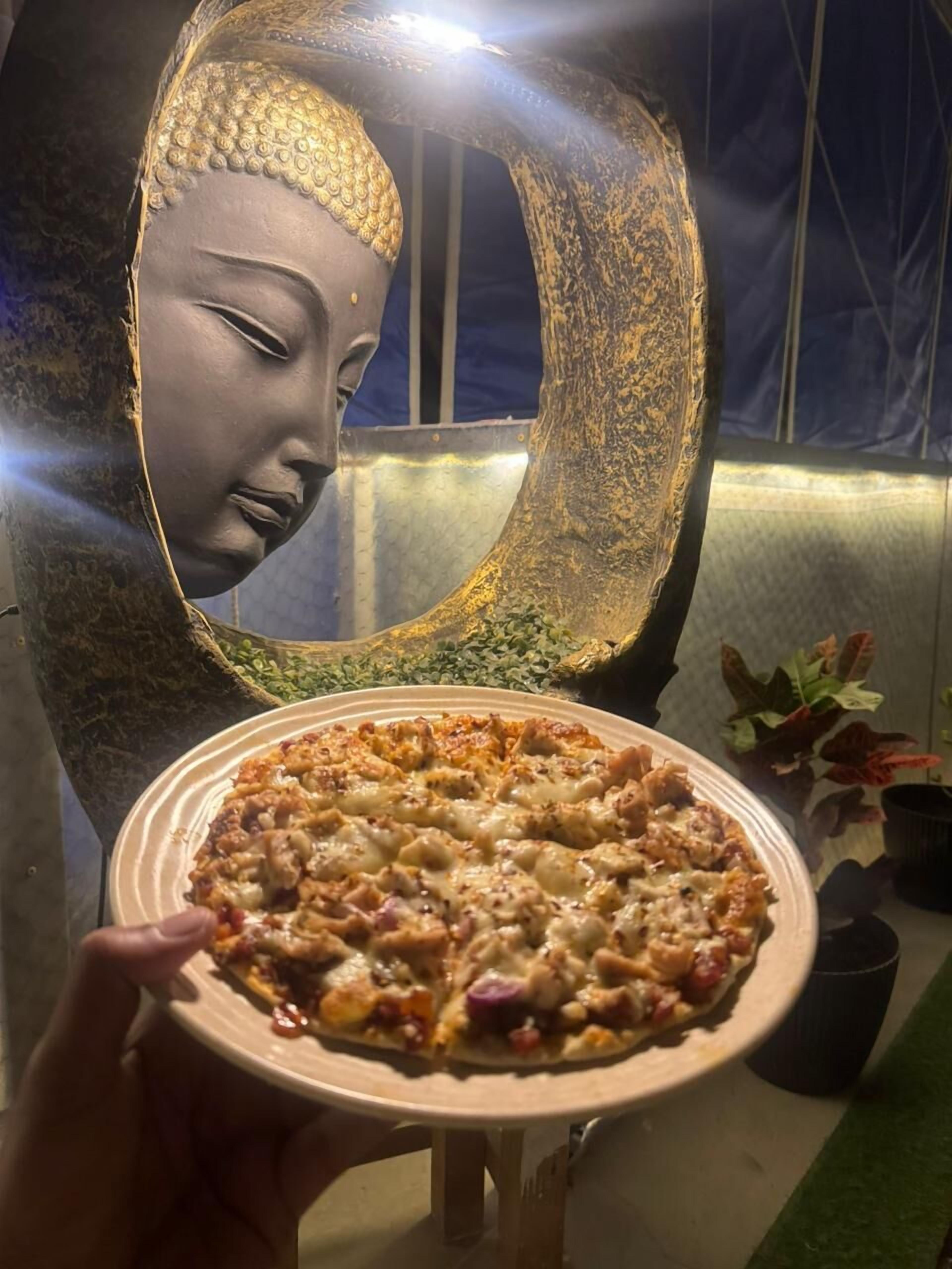 Una persona sosteniendo una pizza, con una estatua de Buda al fondo.