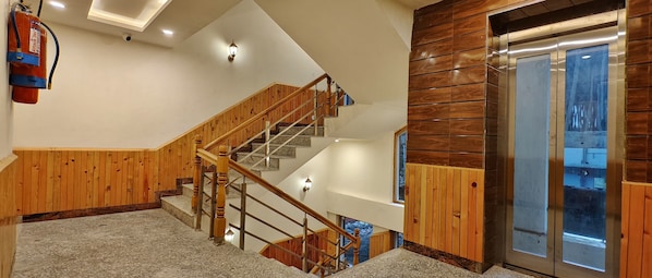 Una escalera con barandillas de madera y peldaños de granito, que conduce a un rellano con un muro de madera y una puerta de ascensor de cristal.