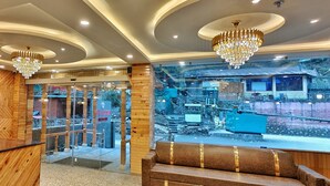 A hotel lobby with wooden paneling, a reception desk, seating area, and a view of the outdoor area through large glass doors. - Hotel Shingar - Sunrise Hotel & Resorts (Bhuntar)