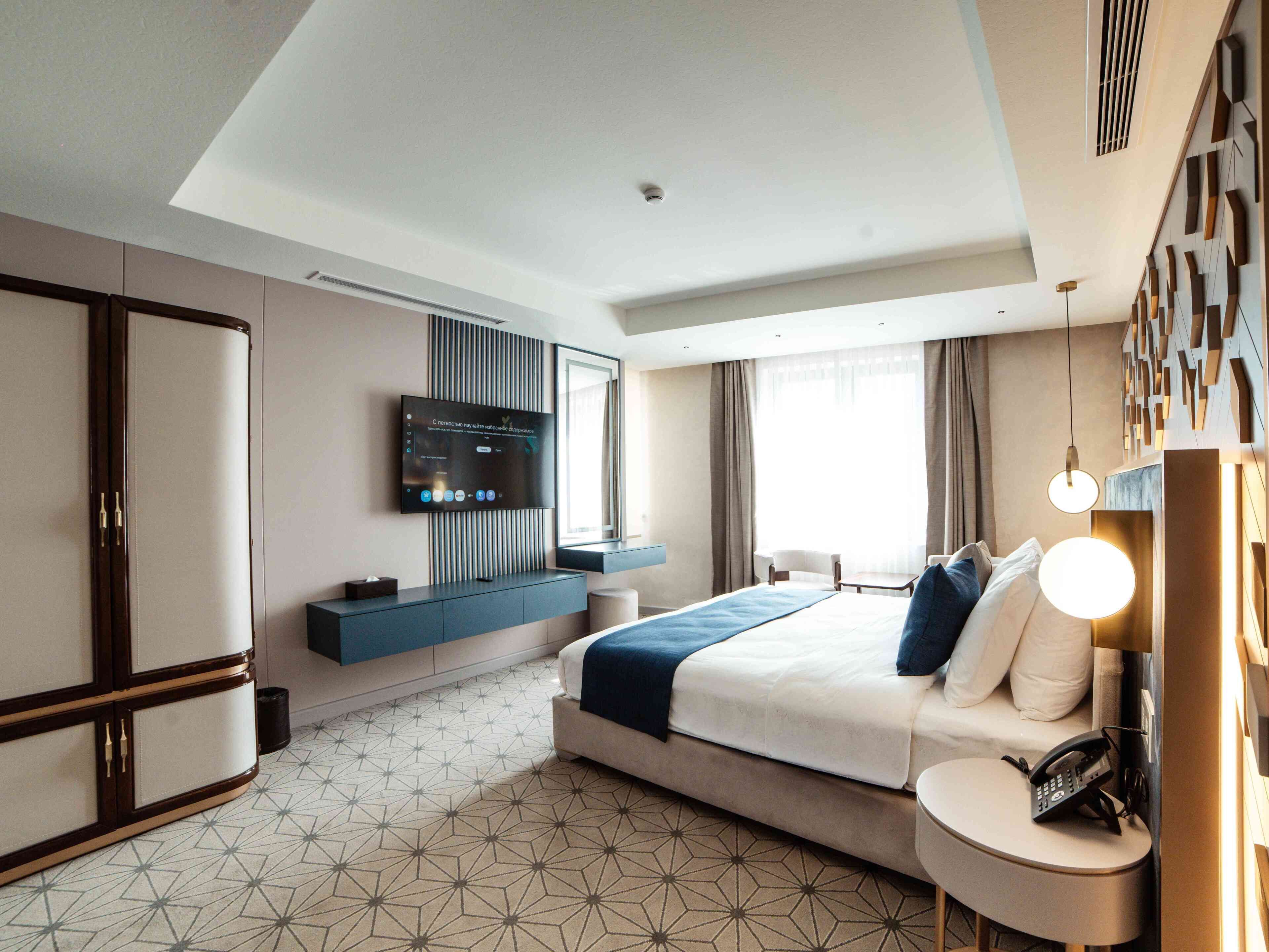 A modern hotel room with a large bed, a TV mounted on the wall, a desk with a lamp, and a wardrobe.