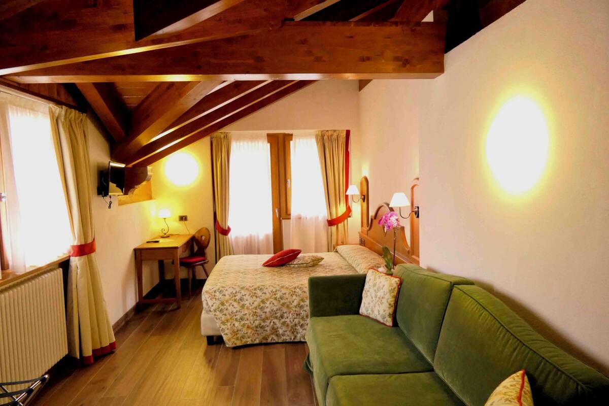 A cozy room with a wooden ceiling, a green sofa, a bed with floral bedding, a desk with a chair, and a radiator.